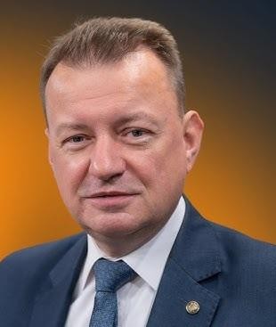 Mariusz Błaszczak przemawiający na konferencji prasowej, z mikrofonami przed sobą i wyraźną gestykulacją.