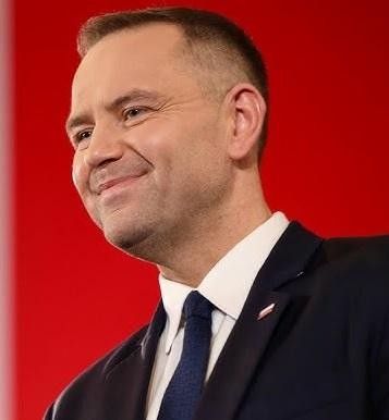 Karol Nawrocki na tle symboli związanych z jego działalnością historyczną i społeczną, takich jak dokumenty, mapy Polski lub elementy upamiętniające Żołnierzy Wyklętych.