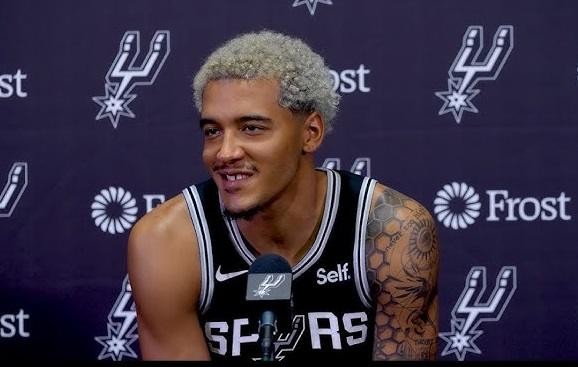 Jeremy Sochan uśmiechnięty i pewny siebie, w koszulce treningowej San Antonio Spurs, trzymający piłkę do koszykówki.
