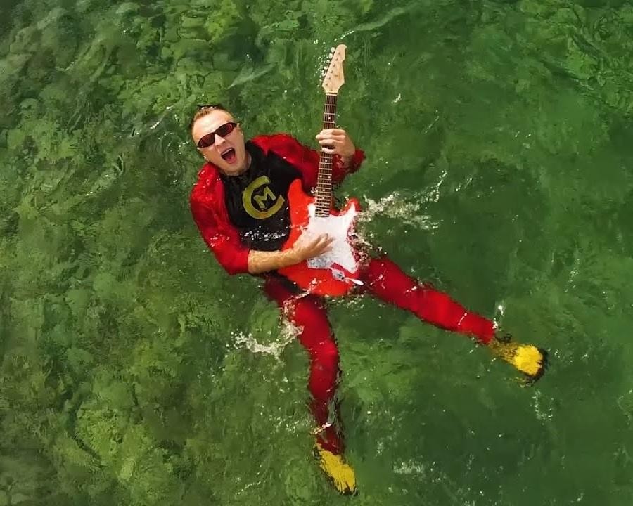 Czadoman w zbliżeniu na scenie, w trakcie występu, z mikrofonem w ręku, w kolorowym stroju scenicznym, z rozmytym tłem świateł estradowych i publiczności.