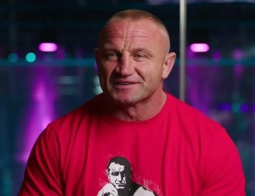 Mariusz Pudzianowski podczas zawodów strongman, z ogromnym wysiłkiem podnosi ciężar, ukazując swoją siłę i determinację, w otoczeniu publiczności.